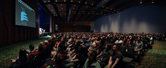 Image of a large audience at a tech conference watching the keynote with live captions above the presentation slides.